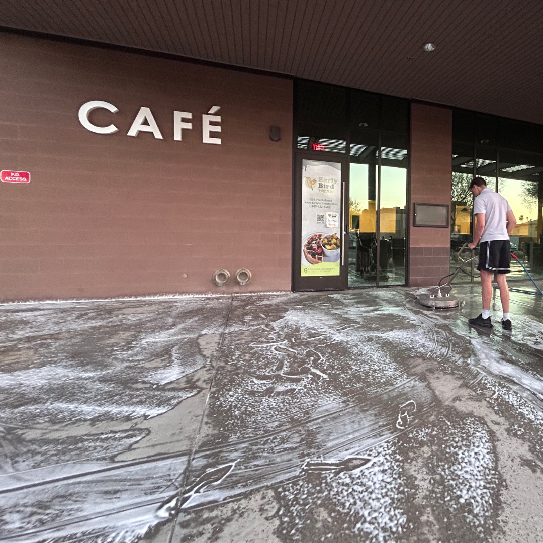 Man using a pressure washer to clean the exterior concrete surface in front of a café, with soap suds visible and café signage in the background.